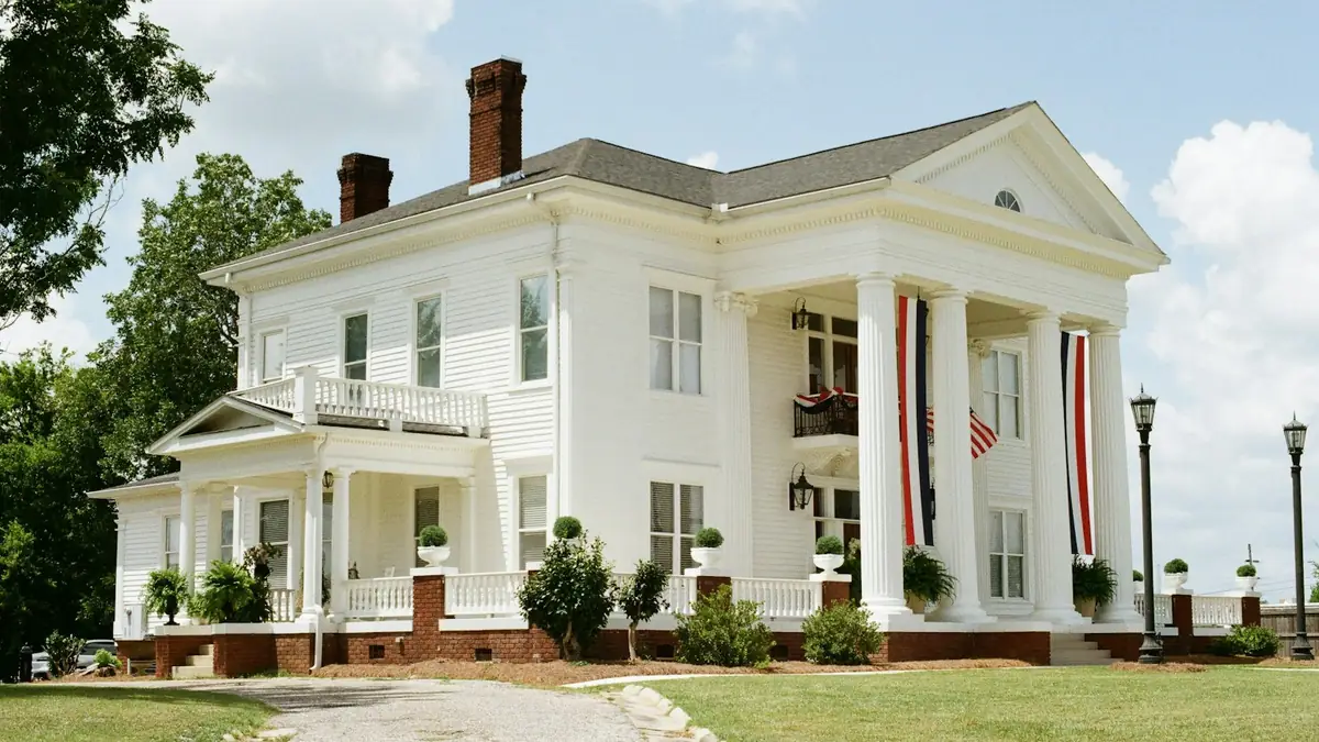 A white two-story house with a columned porch and red-white-and-blue bunting on a lawn - Leak Detection in Petersburg
